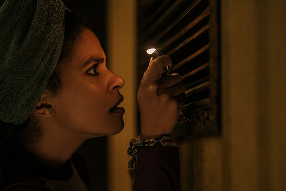 A woman in profile lights a match near a dark window shutter in low light.