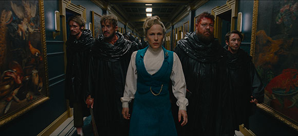 A woman in a blue dress stands in a dark hallway surrounded by men in black, textured robes.