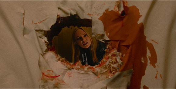A blonde woman peers through a jagged, blood-stained hole in a white sheet.