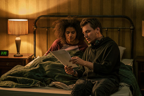A man and a woman sit in a dimly lit bedroom, looking at a piece of paper together.