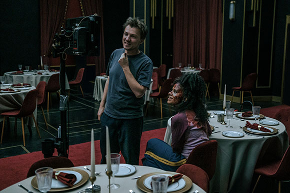 A man in a grey t-shirt stands by a camera while a woman with facial makeup sits at a set table.