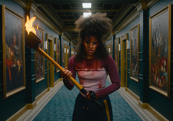 A bloodied woman stands in a hallway holding a large flaming torch.