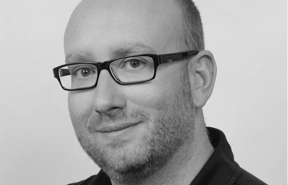 Black and white close-up portrait of a man with short hair, glasses, and light facial stubble.