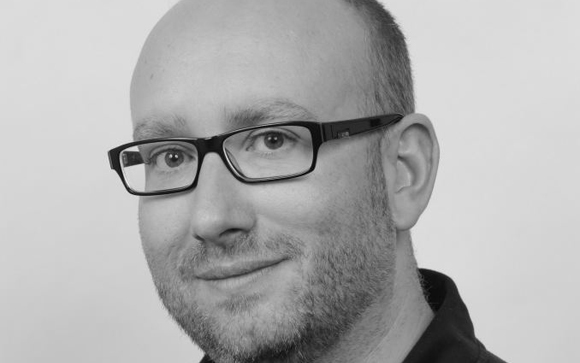Black and white close-up portrait of a man with short hair, glasses, and a beard looking toward the camera.