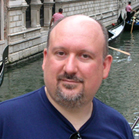 A man with a goatee and dark blue shirt smiling in front of a canal with gondolas.