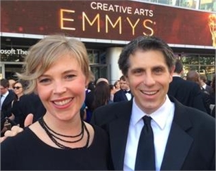 A smiling man in a suit and a woman with blonde hair posing in front of a Creative Arts Emmys backdrop.