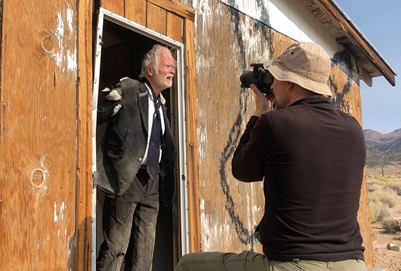 A man in a dark suit stands in a wooden doorway as a photographer takes his picture.