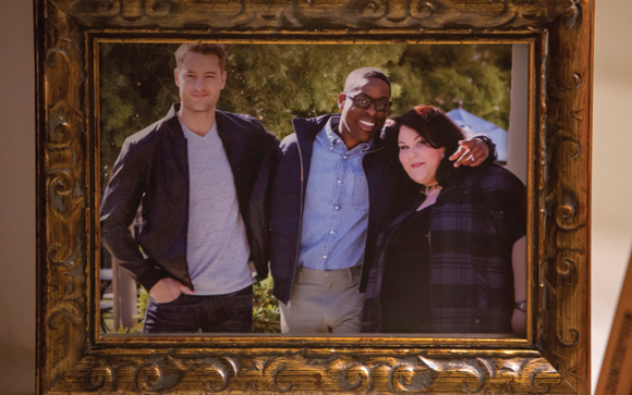 A gold ornate frame holds a photo of a man in a bomber jacket, a man in a blue shirt, and a woman in a plaid jacket.