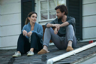 A man and a woman sit side-by-side on a shingled roof in front of a house with dark shutters.