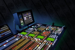 A large audio mixing console with numerous sliders and buttons, next to a computer monitor in a dark room.