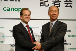 Two men in business suits shake hands in front of a white backdrop with various company logos.