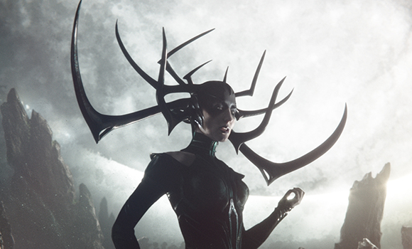 A woman in dark clothing and a large, black, branching antler-like headpiece stands against a cloudy sky.