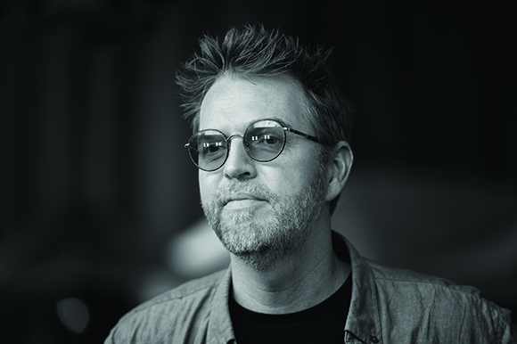 Black and white close-up portrait of a man with short, messy hair and round glasses looking slightly off-camera.
