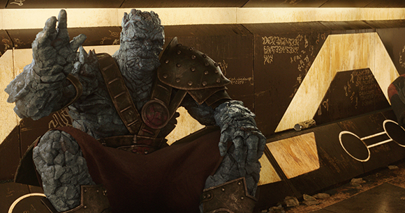 A large, blue, rocky-skinned character wearing bronze armor sits in a metallic environment.