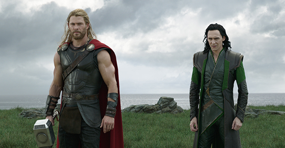 Thor in armor with a red cape and Loki in green and black attire stand in a grassy field under a cloudy sky.