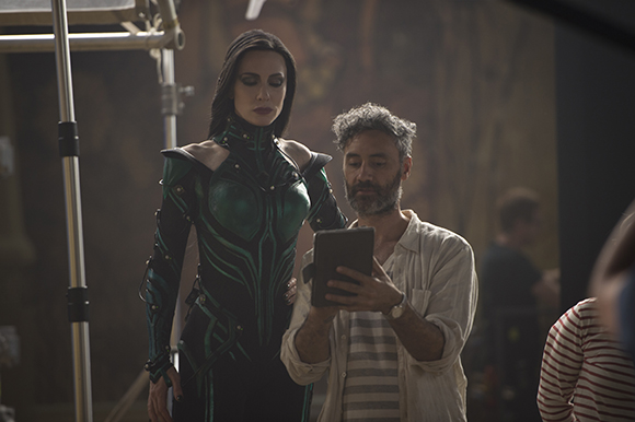 A woman in green and black armor stands beside a man reviewing content on a tablet.