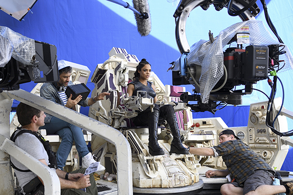A film crew operates cameras and equipment around an actress in a sci-fi cockpit set.