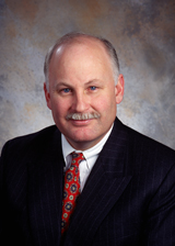 A man with light skin, thinning hair, and a mustache wearing a dark pinstripe suit and red patterned tie.