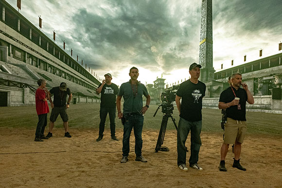 A film crew stands in a large, stone-walled arena under a cloudy sky with a tall monument in the background.