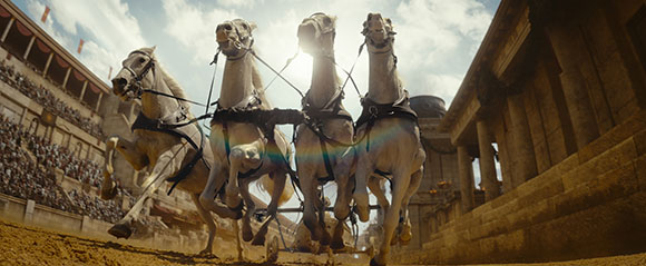 Four white horses galloping forward in a chariot through a sunlit ancient stone arena.