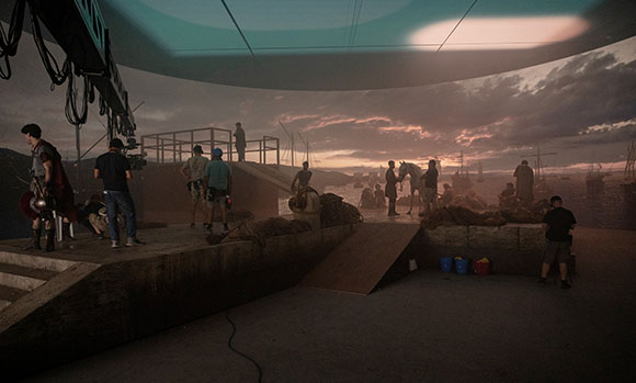 Film crew working on a set with a large sunset sky backdrop and various props and scenery.