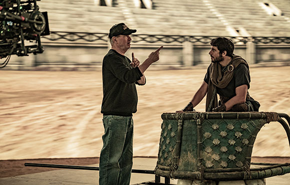 A man in a black shirt and cap points toward an actor dressed in ancient-style clothing sitting in a large prop.