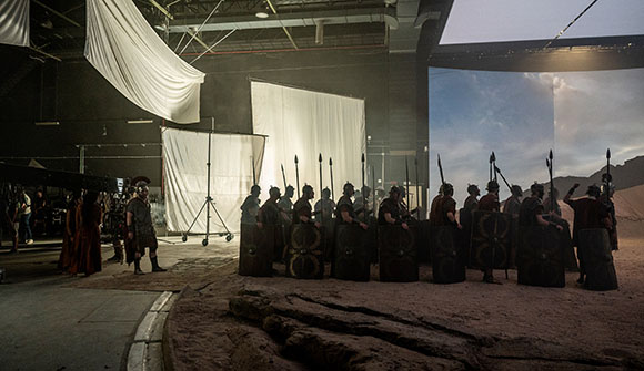 Actors in Roman soldier costumes stand in formation on a large movie studio set with backdrops.