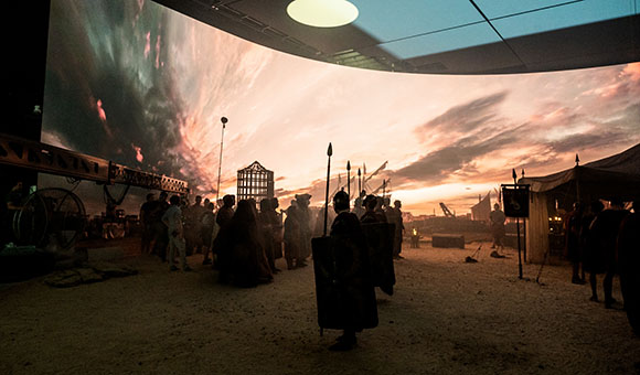 Actors in costume stand on a sandy set in front of a massive curved screen displaying a sunset sky.