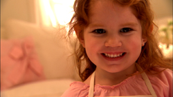 A smiling young girl with curly red hair wearing a pink shirt and a white apron.