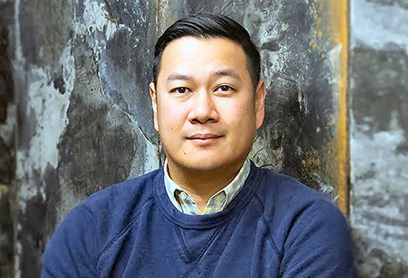 A man with short dark hair wearing a blue crewneck sweater and a light collared shirt.