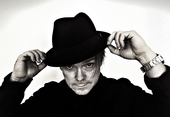 A man wearing a black fedora and dark coat, holding the brim of his hat with both hands.
