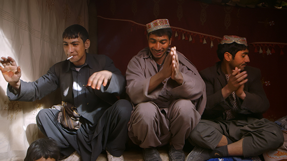 Three men sitting on the floor in a room, two wearing traditional caps and one smoking.