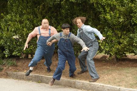 Three men wearing overalls jumping joyfully in front of green bushes.
