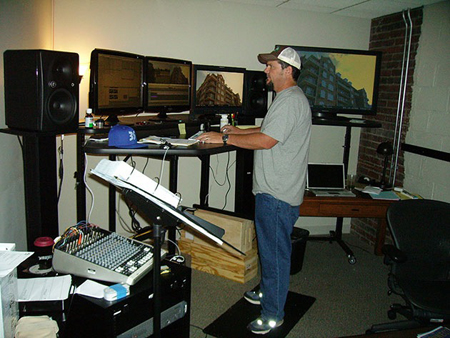 A man in a grey shirt and cap stands at a desk with several computer monitors and a mixing board.