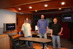 Three men standing in a studio with monitors, mixing equipment, and a wooden ceiling.