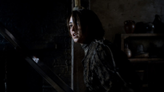 A young person with dark hair looks toward the camera with a startled expression in a dimly lit room.