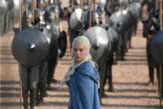 A woman with long blonde hair in a blue cloak stands in front of a formation of soldiers with shields.