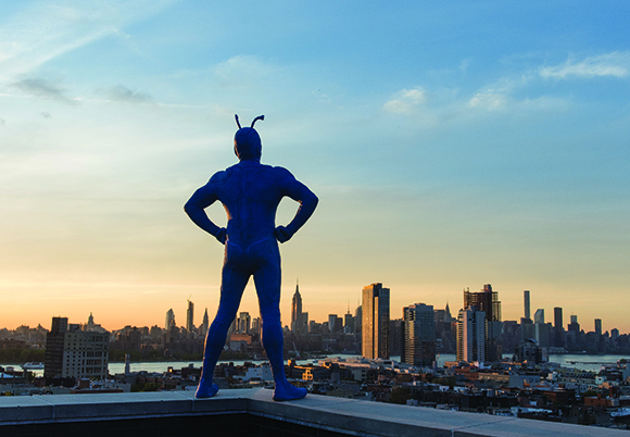 A blue humanoid insect sculpture stands on a rooftop overlooking a city skyline at sunset.