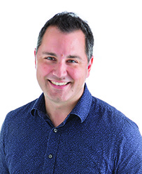 A smiling man with short dark hair wearing a blue button-down patterned shirt against a white background.