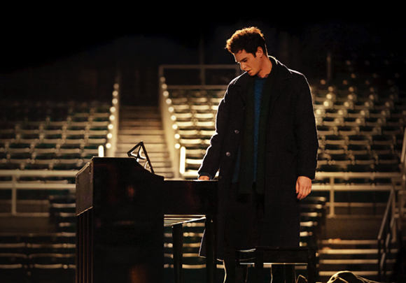 A man in a dark coat and scarf stands by a piano in a dimly lit, empty stadium.