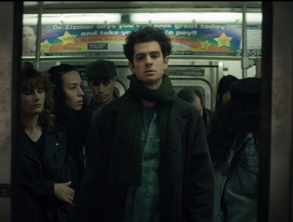 A man with dark curly hair and a green scarf stares forward in a crowded subway car.