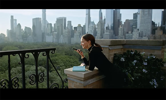 A woman in black leans on a stone balcony railing, looking at her phone with a city skyline in the background.
