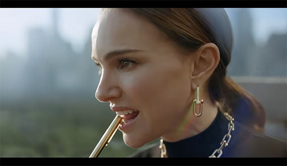 A woman in profile bites the end of a gold lipstick tube against a blurred city background.