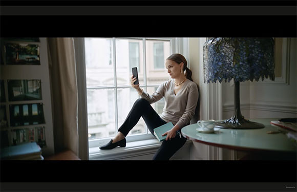 A woman sits on a windowsill holding a smartphone and a blue book near a lamp and a table.