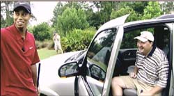 A man in a red shirt stands by a silver car while another man sits in the driver's seat smiling.