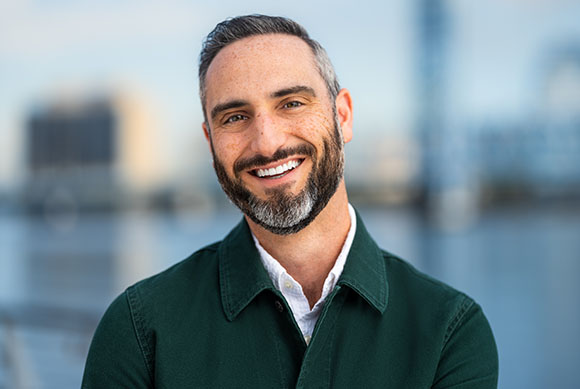 A smiling man with short dark hair and a salt-and-pepper beard wearing a dark green button-down shirt.