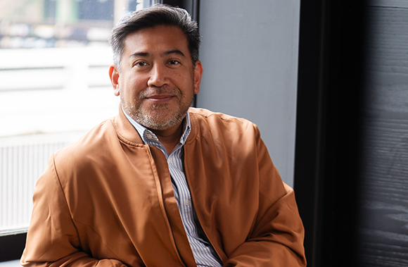 A man with short dark hair and a salt-and-pepper beard wearing a brown bomber jacket.