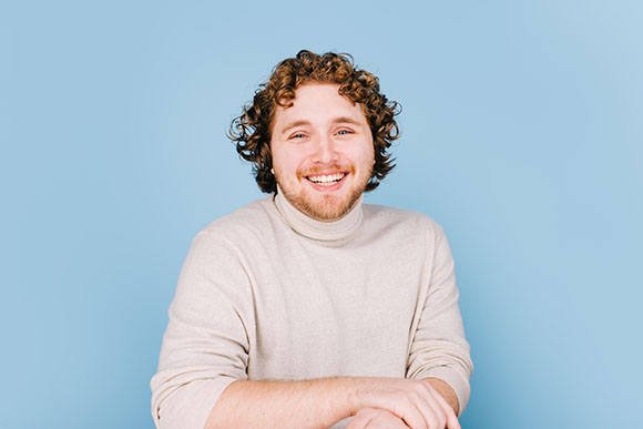 A smiling man with curly brown hair wearing a beige turtleneck against a light blue background.