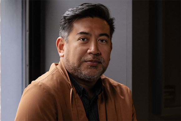 A man with short dark hair and a salt-and-pepper beard wearing a brown jacket.