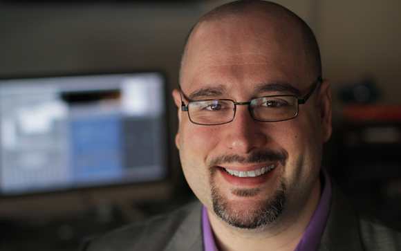 A smiling man with glasses and a goatee, with a computer monitor blurred in the background.